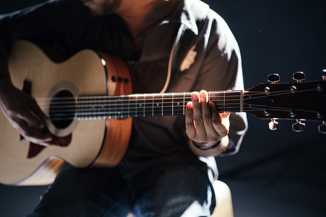 Eine Person spielt Akustikgitarre und konzentriert sich auf ihre Hände, während sie die Saiten zupft. Die Gitarre hat eine natürliche Holzoberfläche und die Szene ist schwach beleuchtet, was einen dramatischen Effekt mit Schatten und Lichtern auf dem Musiker und dem Instrument erzeugt – perfekt für einen Anfänger, der einen Online-Gitarrenkurs belegt.