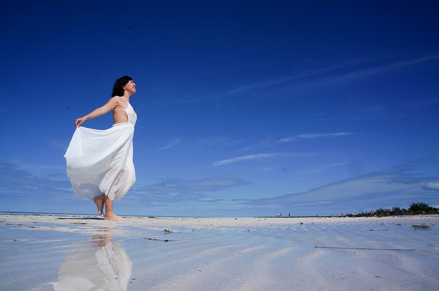 Eine Frau in einem weißen Kleid läuft barfuß an einem Sandstrand entlang, über dem der klare blaue Himmel thront. Sie hält einen Teil ihres Kleides in einer Hand und lässt es in der Brise wehen. Der nasse Sand spiegelt ihr Spiegelbild und trägt zur ruhigen Küstenatmosphäre bei – eine perfekte Szene aus einer Auswandern-Checkliste.