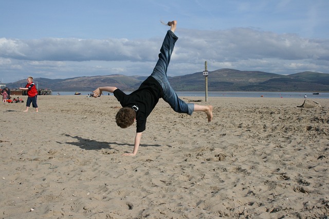 Ein Kind schlägt ein einhändiges Rad an einem Sandstrand mit Bergen und einem bewölkten Himmel im Hintergrund. In der Ferne sieht man andere Kinder und einige Erwachsene, die vielleicht von einem Anfängerhandbuch inspiriert wurden, das sie gefunden haben, und die den Strand nach 30 Tagen Übung genießen.