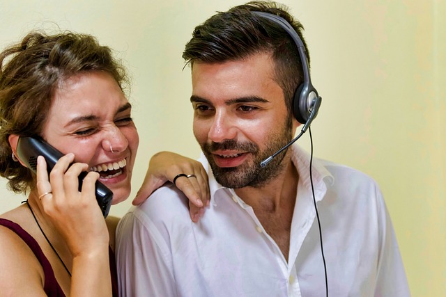 Eine Frau lächelt und telefoniert, während ein Mann neben ihr mit einem Headset in die gleiche Richtung blickt. Sie tragen beide Freizeitkleidung und scheinen das Gespräch zu genießen. Vielleicht besprechen sie ihr Trainingsprogramm der letzten 7 Tage. Der Hintergrund ist neutral.