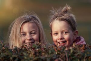 Zwei Kinder mit zerzaustem Haar blicken über die Spitze eines Strauchs und lächeln freudig. Der Hintergrund ist verschwommen, sodass die Aufmerksamkeit auf ihre glücklichen Gesichter gerichtet ist. Die Beleuchtung verleiht der Szene einen warmen, goldenen Schimmer und fängt die pure Essenz ihres Glücks ein, obwohl sie mit ADHS leben.