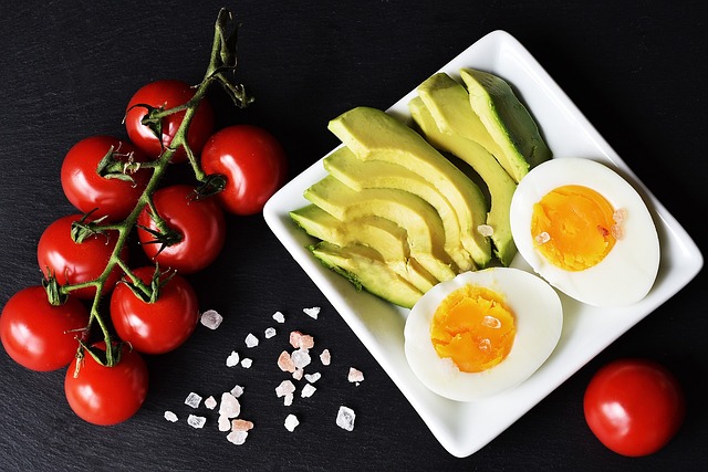 Ein weißer quadratischer Teller auf einer dunklen Oberfläche enthält Avocadoscheiben und ein halbiertes weichgekochtes Ei. Daneben liegen ein Bündel roter Kirschtomaten an der Rebe, ein paar rosa Salzkristalle und eine einzelne lose Tomate. Diese trendige Präsentation ist perfekt für diejenigen, die eine Ketodiät einhalten.