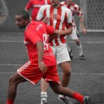 Eine Gruppe Fußballer spielt auf einem Außenfeld Fußball. Im Hintergrund ist ein Tor mit Netz zu sehen. Ein Spieler in einem roten Trikot und weißen Shorts steht im Vordergrund, daneben laufen weitere Spieler in rot-weiß gestreiften Trikots. Der Boden scheint asphaltiert zu sein.