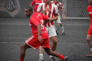 Eine Gruppe Fußballer spielt auf einem Außenfeld Fußball. Im Hintergrund ist ein Tor mit Netz zu sehen. Ein Spieler in einem roten Trikot und weißen Shorts steht im Vordergrund, daneben laufen weitere Spieler in rot-weiß gestreiften Trikots. Der Boden scheint asphaltiert zu sein.