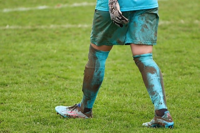 Das Bild zeigt die schlammverkrusteten Beine und den Unterkörper eines Fußballers in hellblauen Shorts und Kniestrümpfen, mit Stollenschuhen und Handschuhen. Der Spieler steht auf einem Rasenfeld mit sichtbaren Schlammspritzern auf seiner Uniform und seinen Beinen, was die intensive Spielvorbereitung dieses Sports verdeutlicht.