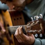 Eine Nahaufnahme einer Person, die Akustikgitarre spielt, perfekt für Anfänger. Der Fokus liegt auf den Händen: Eine drückt die Saiten entlang des Griffbretts nach unten, die andere klimpert in der Nähe des Schalllochs. Mit einem unscharfen Hintergrund, der Gitarre und Hände hervorhebt, ist diese Szene ideal für einen Online-Gitarrenkurs.