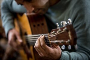 Eine Nahaufnahme einer Person, die Akustikgitarre spielt, perfekt für Anfänger. Der Fokus liegt auf den Händen: Eine drückt die Saiten entlang des Griffbretts nach unten, die andere klimpert in der Nähe des Schalllochs. Mit einem unscharfen Hintergrund, der Gitarre und Hände hervorhebt, ist diese Szene ideal für einen Online-Gitarrenkurs.