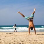 Eine Person macht einen Handstand an einem Sandstrand mit dem Meer im Hintergrund und sieht aus, als hätte sie ihn in nur 30 Tagen gemeistert. Eine andere Person geht mit einem Surfbrett am Ufer entlang. Der Himmel ist klar und blau.