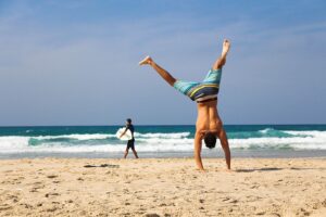 Eine Person macht einen Handstand an einem Sandstrand mit dem Meer im Hintergrund und sieht aus, als hätte sie ihn in nur 30 Tagen gemeistert. Eine andere Person geht mit einem Surfbrett am Ufer entlang. Der Himmel ist klar und blau.