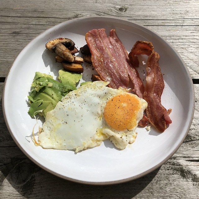 Auf einem Holztisch steht ein trendiger Teller mit Frühstücksgerichten. Er enthält ein Spiegelei, zwei Streifen knusprigen Speck, Avocadoscheiben und sautierte Pilze. Das Ei ist mit schwarzem Pfeffer bestreut.