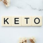 Scrabble-Steine mit der Schreibweise „KETO“ auf einer weißen Marmoroberfläche, umgeben von Büscheln getrockneter weißer Blumen in der oberen linken und unteren rechten Ecke, die den steigenden Trend der Ketodiät im veganen Lebensstil hervorheben.