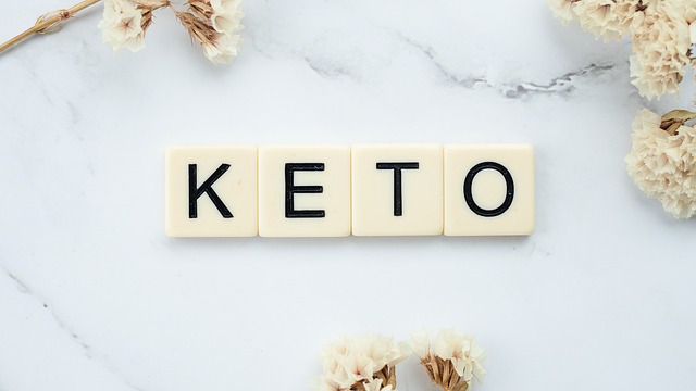 Scrabble-Steine mit der Schreibweise „KETO“ auf einer weißen Marmoroberfläche, umgeben von Büscheln getrockneter weißer Blumen in der oberen linken und unteren rechten Ecke, die den steigenden Trend der Ketodiät im veganen Lebensstil hervorheben.