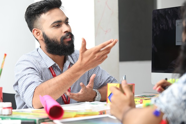 Ein Mann mit Bart und kurzen Haaren gestikuliert mit der Hand, während er mit jemandem außerhalb des Bildes spricht. Er sitzt an einem Schreibtisch voller bunter Schreibwaren und einem offenen Laptop und spricht deutlich, als würde er etwas Wichtiges erklären.