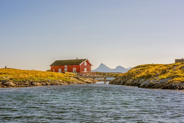 Eine ruhige Küstenszene zeigt eine einsame rote Holzhütte am Wasser, umgeben von gelbgrünen Grasfeldern. Im Hintergrund erheben sich ferne, sonnenbeschienene Berge vor einem klaren blauen Himmel. Das ruhige Wasser im Vordergrund spiegelt die friedliche Atmosphäre wider – ideal, um Ihr Norwegisch in einem Online-Kurs zu üben.
