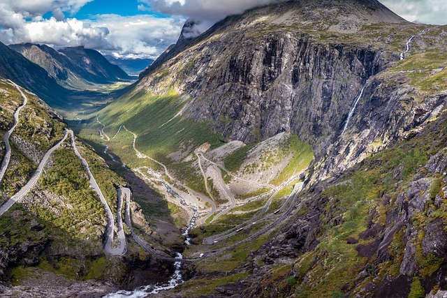 Eine kurvenreiche Straße schlängelt sich durch eine bergige Landschaft mit steilen Klippen, üppiger grüner Vegetation und einem rauschenden Wasserfall. Der Himmel ist teilweise bewölkt und wirft Schatten auf das Gelände. In der Ferne ist ein Tal zu sehen – eine Szene, die sich perfekt für Ihren nächsten Norwegisch-Onlinekurs eignet.