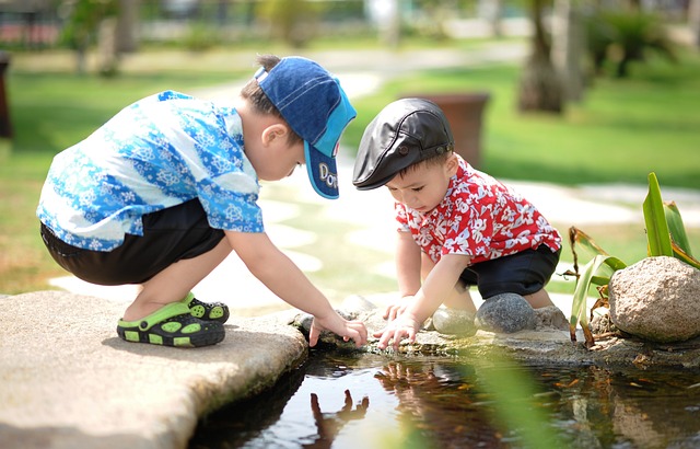 Zwei kleine Kinder, eines mit blauer Mütze und geblümtem Hemd, das andere mit schwarzer Mütze und rotem geblümtem Hemd, hocken neben einem seichten Wasserbecken in einem Park. Sie berühren neugierig das Wasser, umgeben von Grün und Sonnenlicht. Ihr Spiel ist ein friedlicher Moment inmitten ihrer gemeinsamen Erfahrung mit ADHS.