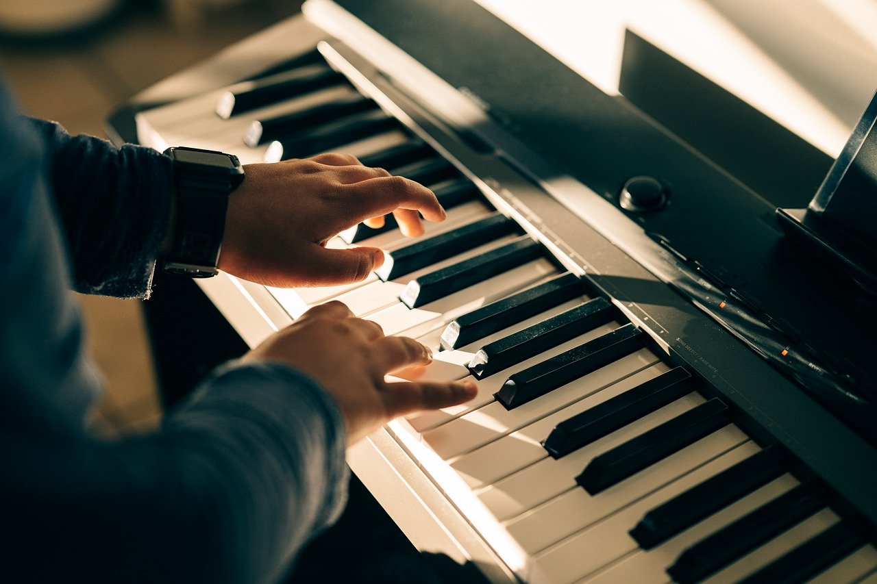 Nahaufnahme einer Person, die während eines Online-Klavierunterrichts auf einem digitalen Keyboard spielt. Die Hände der Person befinden sich über den Tasten, wobei sich die linke Hand auf die tieferen Töne und die rechte Hand auf die höheren Töne konzentriert. Die Person trägt ein dunkles Langarmhemd und eine Uhr am linken Handgelenk.