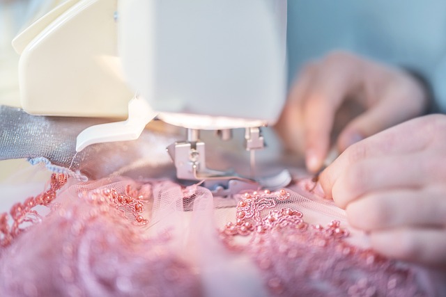 Nahaufnahme von Händen, die einen rosa, aufwendig bestickten Stoff durch eine Nähmaschine führen. Der Fokus liegt auf Nadel und Stoff, wodurch die detaillierte Näharbeit im Gange zur Geltung kommt.