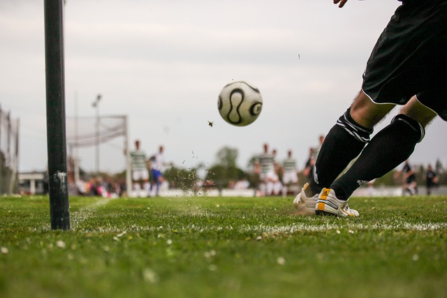 Nahaufnahme eines Fußballers in schwarz-weißer Uniform, der einen kraftvollen Tritt gegen einen Ball ausführt. Der Ball befindet sich in der Luft, im Hintergrund sind die Spieler unscharf zu sehen und ein Torpfosten, was auf ein spannendes Spiel schließen lässt. Durch den Tritt werden Gras und Dreck aufgewirbelt.