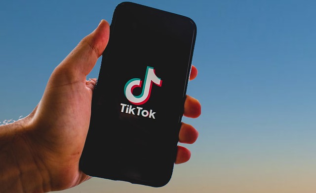 Eine Hand hält ein Smartphone, auf dessen Bildschirm das TikTok-Logo angezeigt wird, das Möglichkeiten zur Monetarisierung von TikTok hervorhebt. Der Hintergrund zeigt einen klaren Himmel mit einem Farbverlauf von Blau oben zu helleren Tönen unten, was entweder einen Sonnenaufgang oder einen Sonnenuntergang suggeriert.