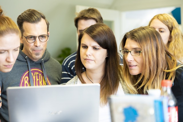 Eine gemischte Gruppe von fünf Erwachsenen sitzt dicht um einen Laptop herum und schaut konzentriert auf den Bildschirm. Sie scheinen gemeinsam an etwas Wichtigem oder Spannendem zu arbeiten, möglicherweise im Zusammenhang mit Seminaren zur Persönlichkeitsentwicklung. Die Umgebung sieht aus wie ein Büro oder ein Gemeinschaftsarbeitsplatz.