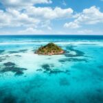 Luftaufnahme einer kleinen, üppig grünen Insel, umgeben von leuchtend türkisfarbenem Ozeanwasser. Das Wasser ist mit dunkleren Flecken von Korallenriffen übersät, und flauschige weiße Wolken schweben darüber in einem strahlend blauen Himmel – wahrlich ein Geheimtipp für jeden, der eine Auswanderung ins Paradies erwägt.