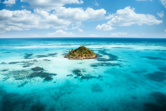 Luftaufnahme einer kleinen, üppig grünen Insel, umgeben von leuchtend türkisfarbenem Ozeanwasser. Das Wasser ist mit dunkleren Flecken von Korallenriffen übersät, und flauschige weiße Wolken schweben darüber in einem strahlend blauen Himmel – wahrlich ein Geheimtipp für jeden, der eine Auswanderung ins Paradies erwägt.