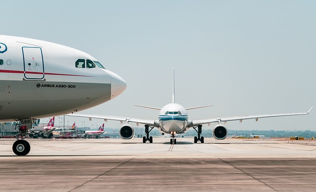 Zwei Verkehrsflugzeuge stehen einander gegenüber auf dem Rollfeld eines Flughafens. Das Flugzeug links ist ein Airbus A330-300, der sich wahrscheinlich auf einen Business Class-Flug vorbereitet. Im Hintergrund in der Nähe des Terminals parken mehrere andere Flugzeuge. Der Himmel ist klar und es scheint Tag zu sein.