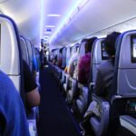 Innenansicht einer Flugzeugkabine, die einen luxuriösen Business Class-Bereich mit in Reihen sitzenden Passagieren zeigt. An der Rückseite jedes Sitzes sind individuelle Bildschirme angebracht, und einige Passagiere verwenden ihre persönlichen elektronischen Geräte. Die Kabine ist mit blauem Umgebungslicht beleuchtet.