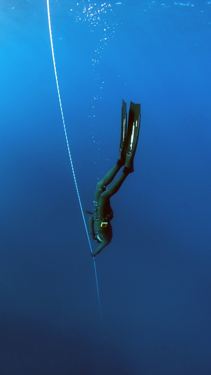 Ein Taucher in einem dunklen Neoprenanzug und mit Flossen taucht senkrecht in tiefblaues Wasser ab und hält sich dabei an einem Führungsseil fest. Luftblasen schweben über ihm, während er tiefer schwimmt und in dieser ruhigen Unterwasserumgebung die Kunst des Apnoetauchens erlernt.