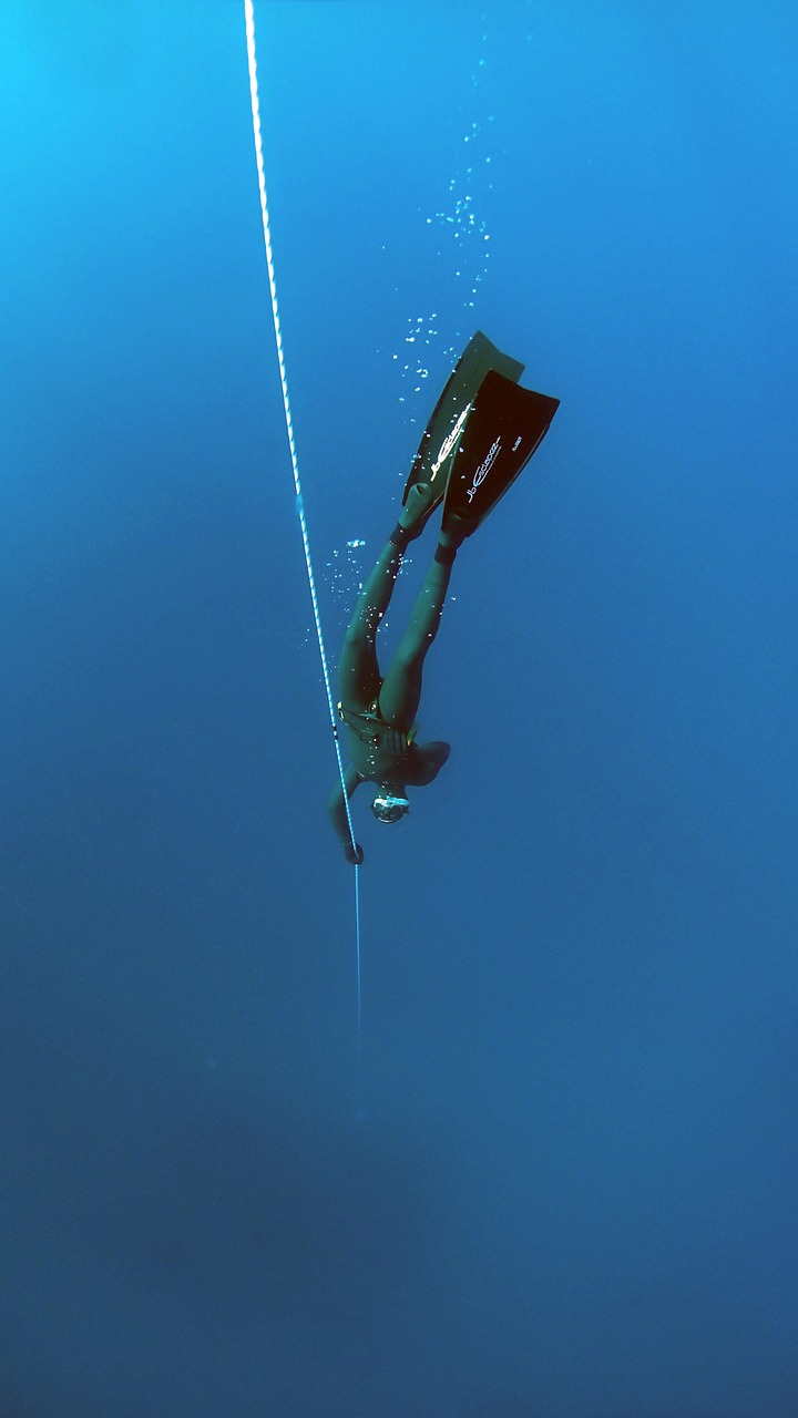 Ein mit Flossen und Schnorchel ausgerüsteter Taucher taucht in das tiefblaue Wasser ab, während er sich an einem Führungsseil festhält. Blasen ziehen hinter ihm her, während er während seines Apnoetauchen-Lernkurses durch die ruhigen, klaren Tiefen navigiert. Der Hintergrund zeigt eine ausgedehnte Unterwasseransicht, die die Einsamkeit des Tauchers betont.