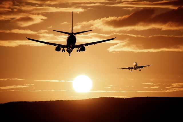 Zwei Flugzeuge sind in der Luft zu sehen, wie sie vor dem Hintergrund eines goldenen Sonnenuntergangs zur Landung ansetzen. Die Sonne steht tief am Horizont und wirft einen orangefarbenen Schein, während vereinzelte Wolken die dramatische Szene verstärken. Die Silhouetten der Flugzeuge sind deutlich zu erkennen – Luxusreisen im Entstehen.